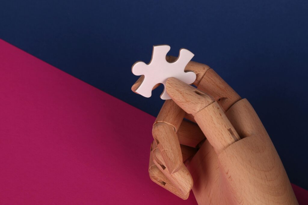 A wooden hand model holding a white jigsaw puzzle piece against a vibrant blue and pink backdrop.